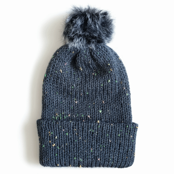 A dark teal knit beanie with colorful specks and a fluffy gray faux fur pom, displayed on a white background.