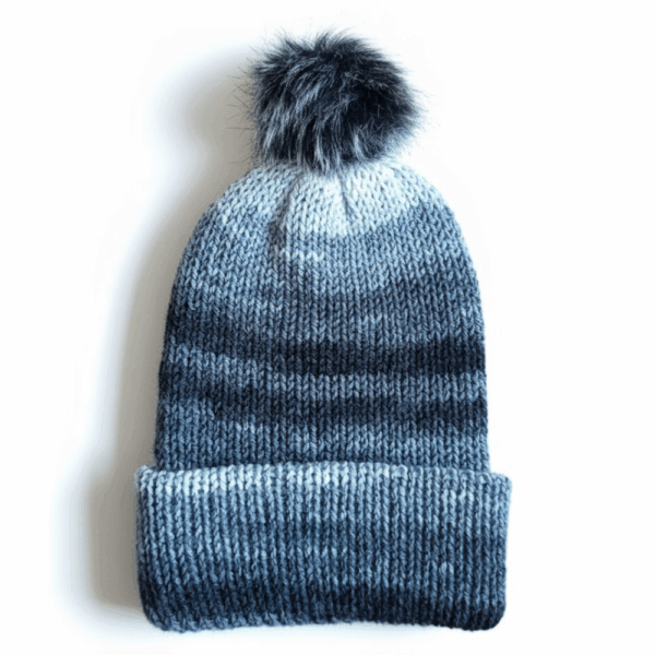 A gradient blue knit beanie with a dark gray faux fur pom on top, displayed on a white background
