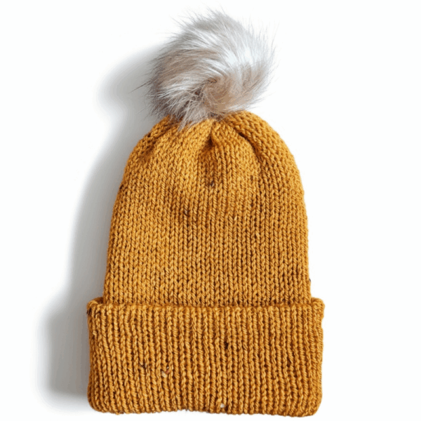 A mustard-yellow knit beanie with a beige faux fur pom on top, displayed on a white background